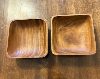 May include: Two square wooden bowls with rounded corners. The bowls are made of a light brown wood and have a smooth, polished finish. The bowls are empty and sit on a wooden surface.