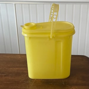 Può includere: Un contenitore rettangolare giallo brillante con coperchio e manico. Il contenitore è in plastica e poggia su una superficie di legno. Il manico è attaccato al coperchio ed è in posizione verticale.