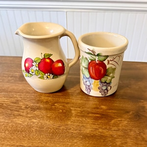 Puede incluir: Un juego de jarra y portautensilios de cerámica color crema. La jarra tiene un asa y está decorada con manzanas rojas y margaritas blancas. El portautensilios presenta una manzana roja y uvas. Ambas piezas tienen un estilo rústico pintado a mano.