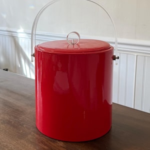 Puede incluir: Una cubitera roja brillante con un asa de acrílico transparente. La cubeta cilíndrica tiene una tapa roja a juego y un pequeño anillo de metal en la parte superior. El asa está sujeta con pequeñas fijaciones metálicas. La cubitera está sobre una superficie de madera.