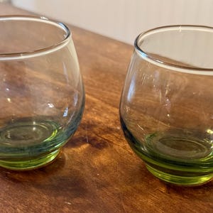 May include: Two clear, stemless drinking glasses with a rounded shape. The base of each glass has a green tint, and the rest of the glass is transparent. The glasses are sitting on a wooden surface.