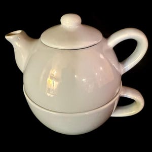 May include: A white ceramic tea set for one, featuring a rounded teapot with a matching lid and a curved handle, stacked securely on top of a matching tea cup with a side handle. The entire piece has a smooth, glossy finish and a solid white colour.