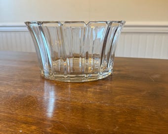 May include: A clear glass bowl with a flared, ribbed design. The bowl sits on a wooden surface, reflecting the light. The bowl's edges have a scalloped shape, adding to its decorative appeal. The bowl is empty and appears to be in good condition.
