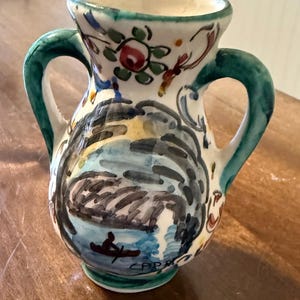 Puede incluir: Un pequeño jarrón de cerámica pintado a mano con dos asas verdes. El jarrón presenta una escena de paisaje con una persona en un bote y diseños florales y abstractos. El jarrón es blanco con detalles en azul, verde, rojo y negro.