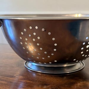 May include: A copper-toned stainless steel colander. This kitchen utensil has a bowl shape, small drainage holes, and two handles. It rests on a circular base. Ideal for draining liquids from food, it measures approximately 25 cm in diameter.