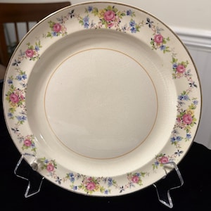 Puede incluir: Plato color crema con borde dorado y borde floral con rosas rosas, flores azules y amarillas y hojas verdes. El plato se exhibe sobre un soporte transparente, mostrando su diseño vintage.