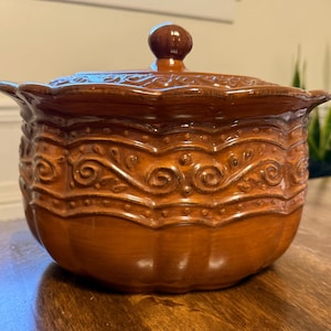 May include: A brown ceramic casserole dish with a lid. The dish has a decorative, embossed floral pattern around the sides and a small knob on the lid. The dish is sitting on a wooden surface.