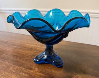 Könnte beinhalten: Eine leuchtend blaue Glas-Fußschale mit gewelltem Rand und dunklem Rand. Die Schale wird von einem ausgestellten Sockel getragen. Die Schale wird wahrscheinlich zum Servieren von Obst oder als dekoratives Stück verwendet.