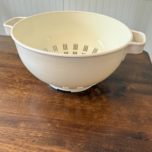 May include: A beige plastic colander with a wide bowl shape and two handles. The colander has small holes in the bottom and sides for draining. It sits on a dark brown wooden surface.