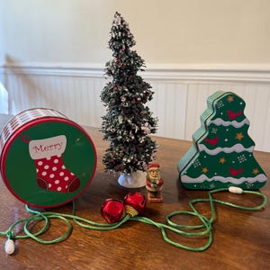 Puede incluir: Una escena navideña festiva con una lata verde y roja con un diseño de calcetín, un árbol de Navidad en miniatura flocado, una lata en forma de árbol y una figura de Papá Noel. Campanas rojas y un cordón verde se suman a la decoración navideña.