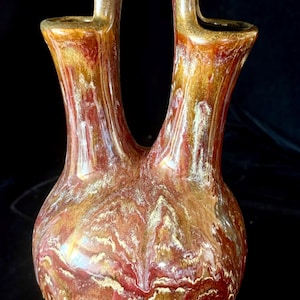 May include: A ceramic double-spout vessel featuring a rounded base and a bridge handle connecting the two openings. The surface displays a marbled glaze in shades of deep red, golden brown, and cream, transitioning to a solid matte olive green base.
