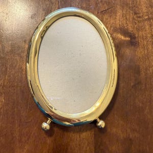 May include: A gold-colored oval picture frame with a cream-colored interior. The frame has a decorative design and small ball feet. The frame is sitting on a wooden surface.