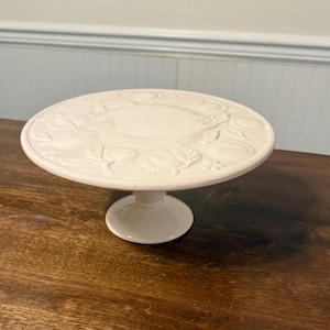 May include: A cream-colored ceramic cake stand with a round top and a pedestal base. The top features a raised fruit and leaf design. The stand is on a dark wood surface.