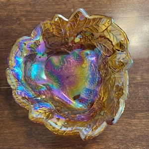 May include: Iridescent glass bowl with a leaf and berry design. The bowl has a scalloped edge and a vibrant colour palette of gold, purple, and teal. The bowl is likely vintage or antique.