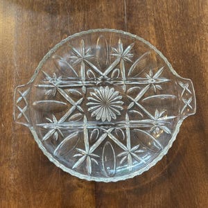 Pode incluir: Prato de servir de vidro transparente com um design de estrela e geométrico. O prato tem uma borda ondulada e duas alças. O centro apresenta um padrão em forma de flor, e o design geral é simétrico. O prato é provavelmente usado para servir aperitivos ou sobremesas.