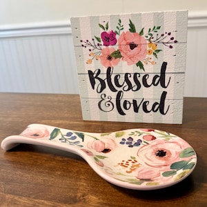 以下が含まれることがあります： ピンク、緑、青の花柄のセラミック製スプーンレスト。スプーンレストは、「Blessed & loved」の文字とフラワーアレンジメントが施された木製のブロックサインの隣にあります。