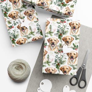 May include: Gift wrap featuring a repeating pattern of smiling yellow Labrador Retrievers, interspersed with green foliage and red berries. The wrapped gifts are tied with twine and adorned with white tags. A spool of twine and scissors are also visible.