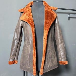 May include: A brown leather jacket with a faux fur lining and trim. The jacket features a classic design with a notched collar, long sleeves, and pockets. The interior and trim are a warm orange color, providing a striking contrast to the brown exterior.