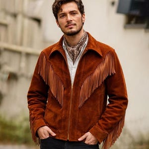 May include: A rust-colored suede jacket featuring long fringe detailing across the chest and down the sleeves. The jacket has a pointed collar, a central gold-toned zipper closure, and is worn over a white button-down shirt with a patterned neck scarf.