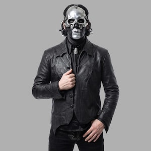 Puede incluir: Una persona con una máscara de calavera plateada y una chaqueta de cuero negra. La chaqueta tiene cierre de botones y cuello con solapas. La persona también lleva una camisa negra y pantalones negros. El fondo es gris liso.