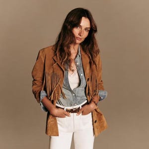 May include: A tan suede jacket featuring long fringe detailing on the chest, a collared neckline, and dark buttons. It is worn over a light blue button-down shirt with a white lace-trimmed camisole underneath, paired with white trousers and a brown belt.