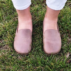 May include: A pair of taupe-colored leather loafers with a rounded toe and white stitching around the sole. The shoes are set on a bed of green grass, with white pants visible above the shoes.