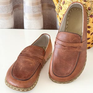 May include: A pair of brown leather loafers featuring a rounded toe, a classic penny slot strap across the vamp, and visible light-coloured stitching along the sole edge. The shoes have a smooth, slightly distressed finish and a light interior lining.