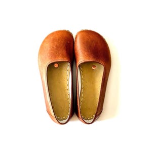 Op de afbeelding: Een paar bruine leren ballerina's. De schoenen hebben een ronde neus en een platte zool. De binnenkant is lichtbruin en de schoenen zijn gestikt met witte draad. De schoenen worden op een witte achtergrond gepresenteerd.