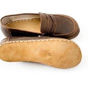 May include: A pair of brown leather loafers featuring a classic penny slot design on the upper and a light tan, textured suede sole. The shoes are constructed with visible, thick white stitching along the perimeter connecting the upper to the sole.