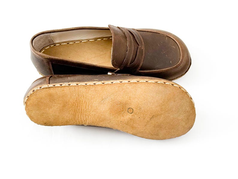 May include: A pair of brown leather loafers featuring a classic penny slot design on the upper and a light tan, textured suede sole. The shoes are constructed with visible, thick white stitching along the perimeter connecting the upper to the sole.