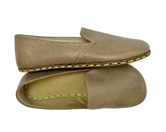 May include: A pair of taupe slip-on shoes featuring a smooth leather upper and a rounded toe. The shoes are constructed with a visible, thick cream-colored whipstitch detail along the sole edge, connecting the upper to the base of the shoe.