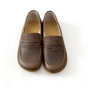 May include: A pair of dark brown leather loafers featuring a classic penny slot design across the vamp, rounded toes, and visible light-colored stitching along the inner collar and the perimeter of the sole, presented on a plain white background.