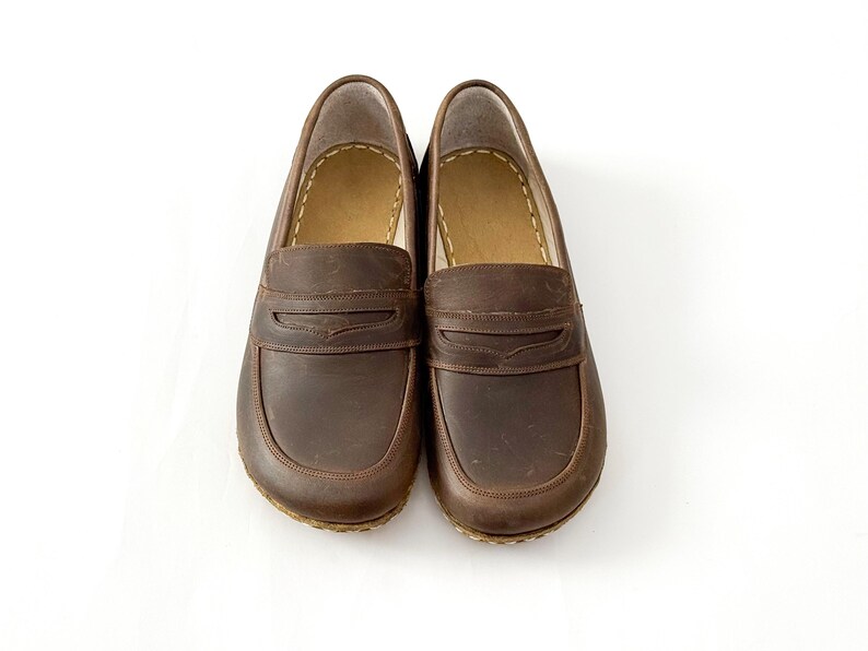 May include: A pair of dark brown leather loafers featuring a classic penny slot design across the vamp, rounded toes, and visible light-colored stitching along the inner collar and the perimeter of the sole, presented on a plain white background.