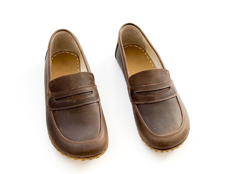 May include: A pair of brown leather loafers featuring a classic penny slot design across the vamp, rounded toes, and visible light-colored stitching along the sole edge, with a tan interior lining and a smooth, matte finish on the exterior leather.