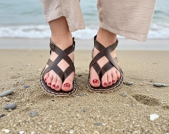 May include: Brown leather sandals with a crisscross design and ankle straps. The sandals have a toe loop and are stitched with white thread. The image shows the sandals on a sandy beach.