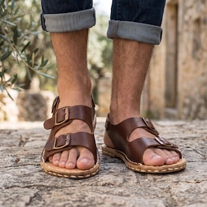 Puede incluir: Un par de sandalias de cuero marrón oscuro con dos correas anchas en la parte superior, cada una asegurada con una hebilla de metal color bronce. Las sandalias tienen un borde de suela cosido de color claro y un diseño abierto con talón.