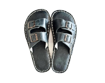 May include: A pair of black sandals featuring two wide straps across the top, each secured with a dark metal buckle. The edges of the soles are finished with prominent white contrast stitching that outlines the entire shape of the footwear.