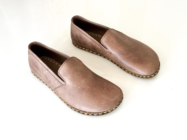 May include: A pair of slip-on shoes crafted from smooth, light brown leather. These shoes feature a rounded toe design, a low-cut collar, and a visible light-coloured whipstitch detail along the outer edge where the upper meets the sole.