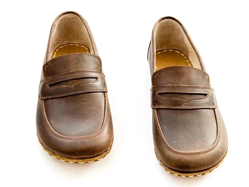 May include: A pair of brown leather penny loafers featuring a smooth, matte finish, a classic strap across the vamp, and visible light-colored stitching along the sole edge. The shoes have a rounded toe design and a tan interior lining.