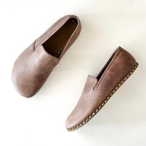May include: A pair of taupe leather slip-on shoes featuring a rounded toe, a low-cut collar, and visible light-coloured whipstitching along the sole edge. The leather has a smooth, matte finish with subtle natural variations in the surface texture.