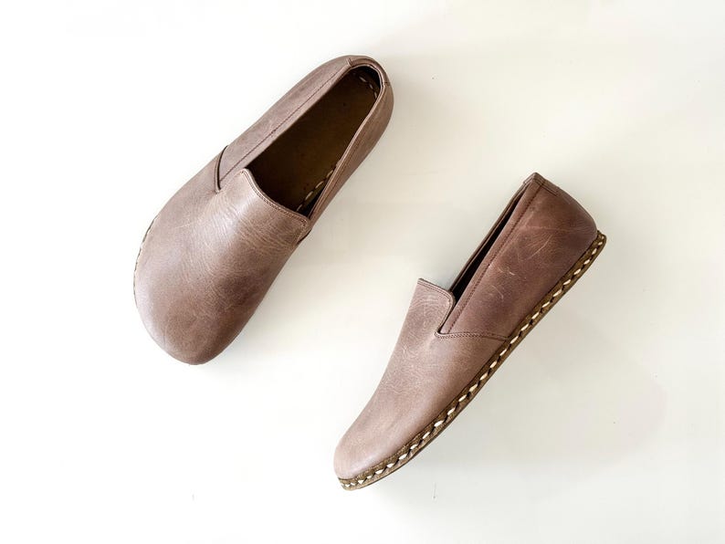 May include: A pair of taupe leather slip-on shoes featuring a rounded toe, a low-cut collar, and visible light-coloured whipstitching along the sole edge. The leather has a smooth, matte finish with subtle natural variations in the surface texture.