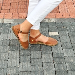 May include: A pair of tan leather flat sandals featuring a closed-toe design, an ankle strap with a small metal buckle, and visible light-colored stitching along the sole edge. The sandals are worn with white trousers against a paved background.