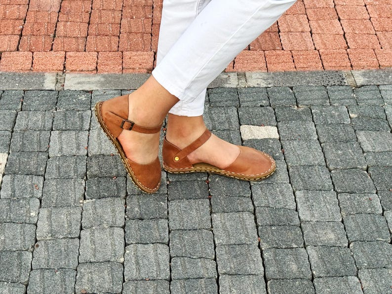 May include: A pair of tan leather flat sandals featuring a closed-toe design, an ankle strap with a small metal buckle, and visible light-colored stitching along the sole edge. The sandals are worn with white trousers against a paved background.