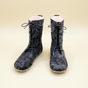 May include: A pair of mid-calf boots featuring a dark gray and black marbled pattern. The boots have a front lace-up closure with thin black laces and a light pink interior lining. The soles are thin and tan, providing a flat base for the footwear.