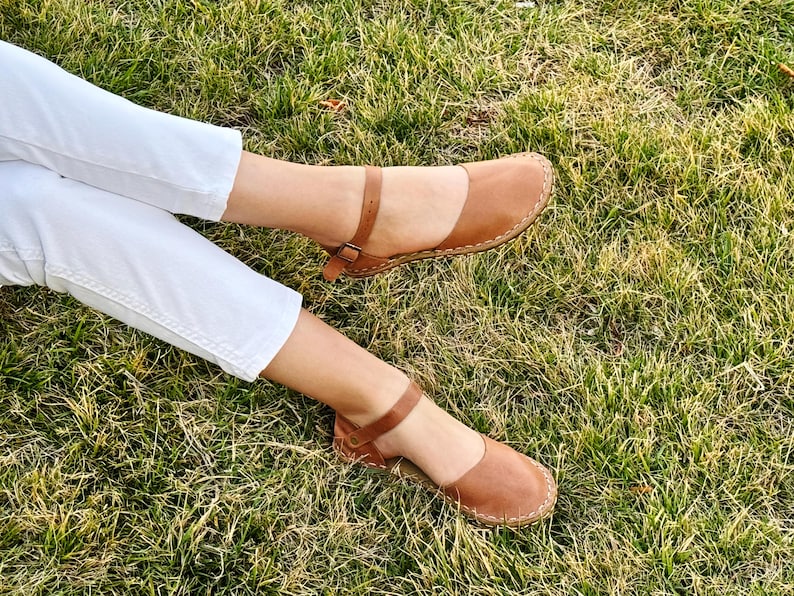 May include: A pair of tan leather sandals featuring a rounded closed toe, an adjustable ankle strap with a small metal buckle, and visible stitching along the sole edge. The footwear is worn with white trousers, showcasing the tan leather construction.