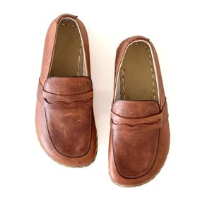 May include: A pair of brown leather loafers featuring a classic penny slot design across the vamp, rounded toes, and visible stitching along the collar and sole edges. The shoes have a smooth, textured finish and a tan interior lining.