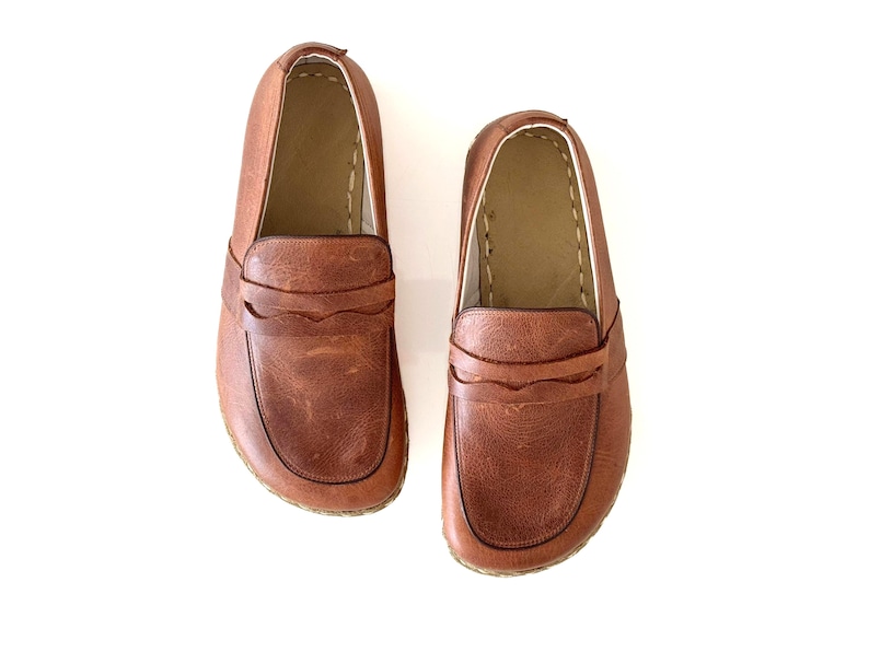 May include: A pair of brown leather loafers featuring a classic penny slot design across the vamp, rounded toes, and visible stitching along the collar and sole edges. The shoes have a smooth, textured finish and a tan interior lining.