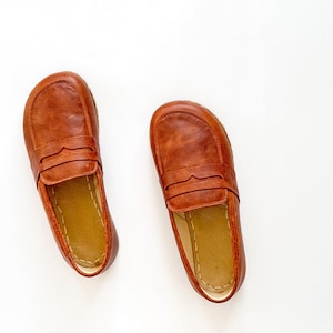 May include: A pair of cognac-colored leather loafers featuring a rounded toe, a classic penny slot strap across the vamp, and visible light-colored stitching along the inner sole edge. The shoes have a smooth finish and a low-profile design.
