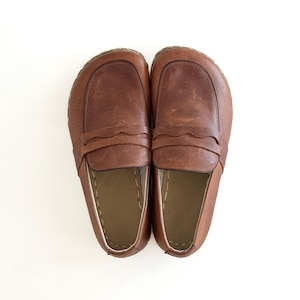 May include: A pair of brown leather loafers featuring a wide, rounded toe box, a classic penny slot strap across the vamp with a scalloped edge, and visible light-coloured stitching along the interior rim of the shoe opening.