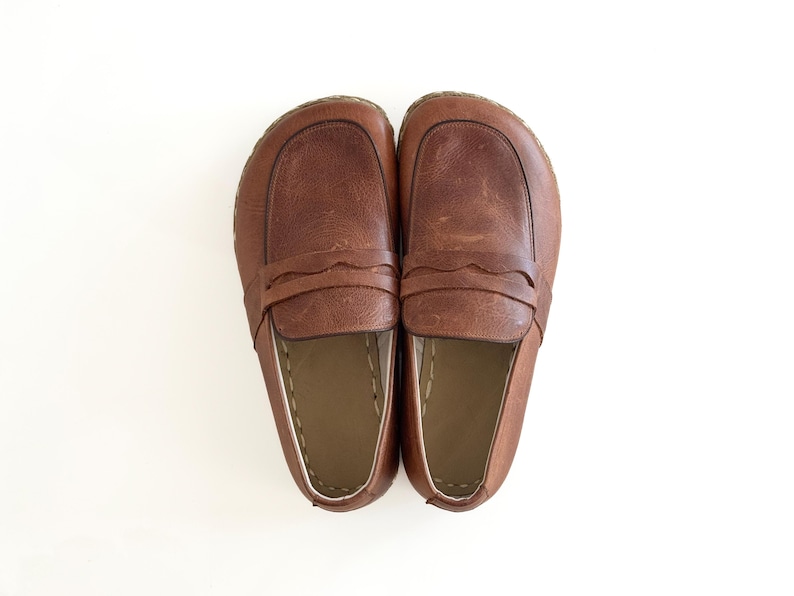 May include: A pair of brown leather loafers featuring a wide, rounded toe box, a classic penny slot strap across the vamp with a scalloped edge, and visible light-coloured stitching along the interior rim of the shoe opening.